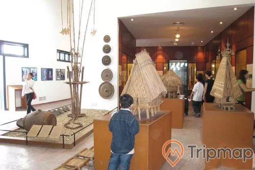 đứa bé đang đứng nhìn mô hình ngôi nhà dân tộc, dụng cụ đồ dùng treo trên tường