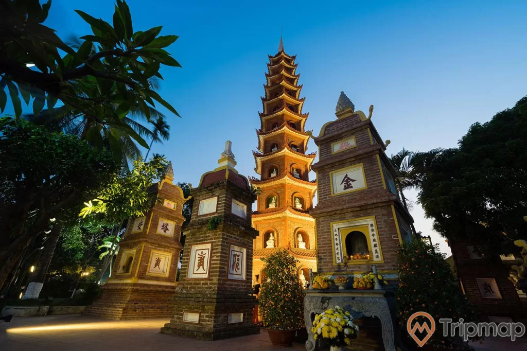 Bảo tháp lục độ đài sen và 3 toà tháp thấp, cây cối và bầu trời xanh tối, 2 chậu cây quất bên cạnh mộ và 1 chậu hoa cúc, đĩa hoa cúc trên bàn đá