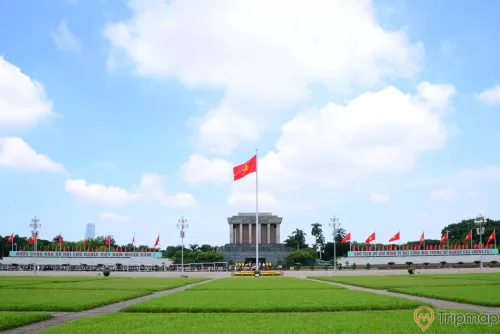 14 lá cờ Việt Nam ở phía xa gần Lăng chủ tịch Hồ Chí Minh, 1 lá cờ lá cờ đỏ sao vàng đang bay phất phới, 4 cây đèn lớn và sân ở cỏ ở trước lăng bác, cây cối mọc um tùm quanh Lăng Bác, bầu trời trong xanh có mây