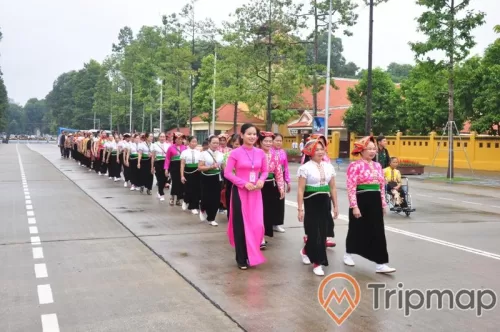 đoàn diễu hành người dân tộc trên đường, một người phụ nữ ngồi xe lăn, hàng cây xanh tươi mọc cạnh nhà mái ngói màu đỏ