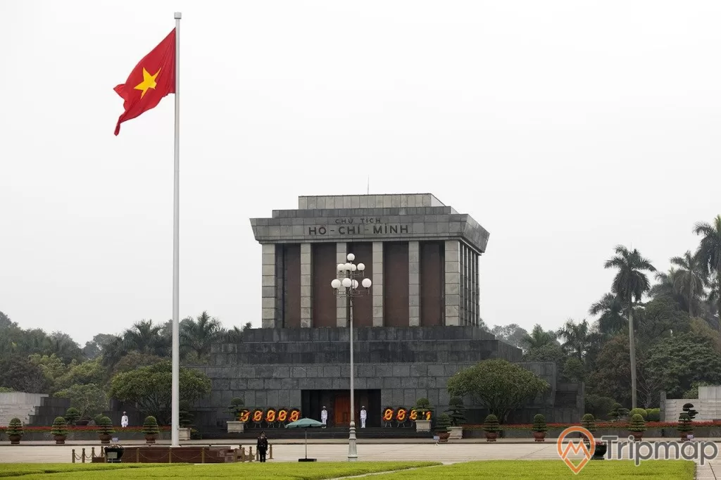 lá cờ đỏ sao vàng Việt Nam đang tung bay, cây cột đèn và cái ô màu xanh lá ở trên sân quảng trường, có nhiều chậu cây cảnh và cây cối mọc cạnh lăng bác, 4 người lính gác đứng bên cạnh lăng bác, bầu trời buổi sáng
