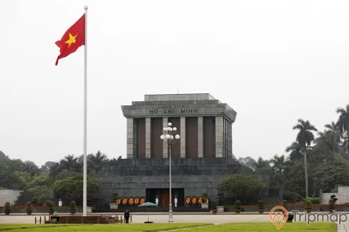 lá cờ đỏ sao vàng Việt Nam đang tung bay, cây cột đèn và cái ô màu xanh lá ở trên sân quảng trường, có nhiều chậu cây cảnh và cây cối mọc cạnh lăng bác, 4 người lính gác đứng bên cạnh lăng bác, bầu trời buổi sáng