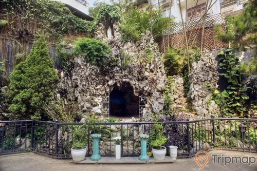 một chiếc hang đá nhân tạo và cây cối mọc trên hang um tùm, trước của hang có chậu cây cảnh nhỏ, trên đỉnh của hang đá có 1 bức tượng đức mẹ