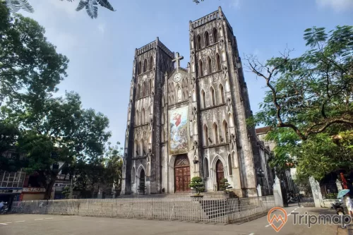 xung quanh nhà thờ lớn hà nội cây cối mọc xanh tươi, có một người cầm ô đứng cạnh chiếc xe máy, nhà thời có hàng rào chắn xung quanh trong sân có 5 chậu cây cảnh